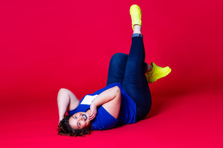 Happy plus size model in casual clothes, fat woman in blue jeans, t-shirt and yellow sneakers on red...