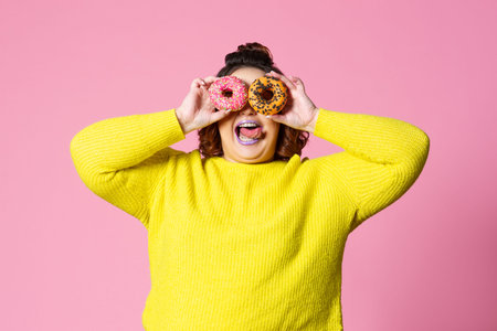 Cheerful plus size model holding donuts against her eyes on pink background, body positive conceptの写真素材