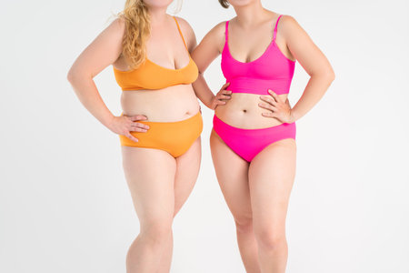 Two overweight women with cellulitis, fat flabby bellies, legs, hands, hips and buttocks on gray studio background, obese female body, liposuction and plastic surgery conceptの写真素材