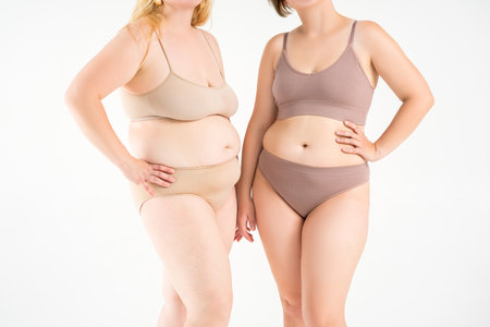 Two overweight women with cellulitis, fat flabby bellies, legs, hands, hips and buttocks on gray studio background, obese female body, liposuction and plastic surgery conceptの写真素材