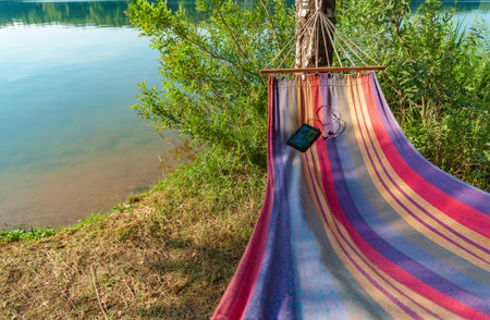 a mobile phone with connected headphones lies in a colored hammock against the backdrop of a lake,...
