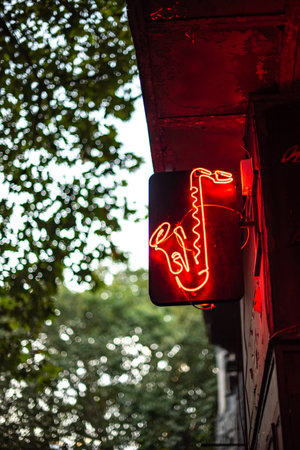 A glowing red neon saxophone sign hanging outside, surrounded by green foliageの写真素材