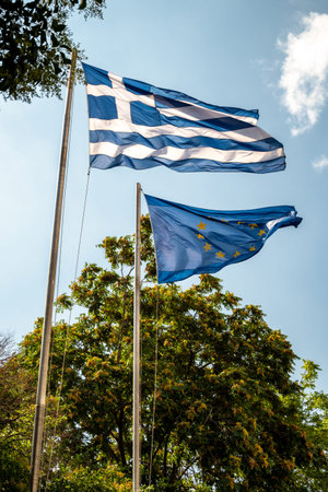 Greek and European Union flags waving on flagpoles against a blue sky.