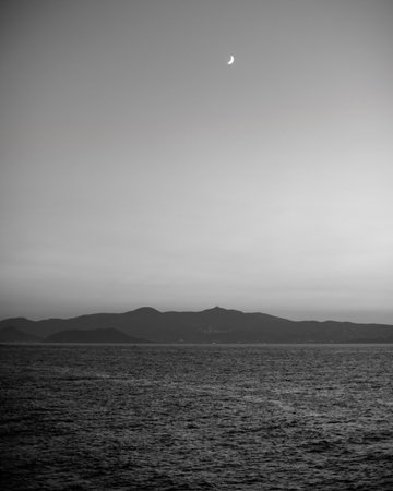 Black and white photo of a crescent moon over a calm sea with distant mountains.の写真素材