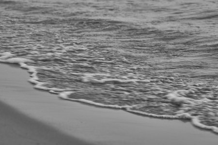 Black and white photo of gentle ocean waves lapping at the sandy shore.の写真素材