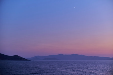Serene seascape at dusk with crescent moon and distant hills.の写真素材