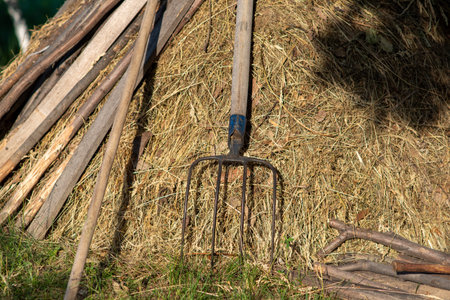Rustic farm pitchfork leaning on a haystack with wooden poles nearby, sunlit rural scene, harvest...
