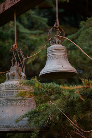 Close-up of two vintage metal bells hanging from rusty wires on an outdoor beam, with evergreen pine branches in the background; rustic church atmosphere.の写真素材