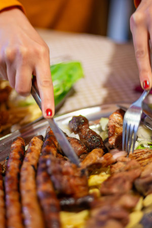 Close-up of hands using fork and knife to cut grilled sausages and mixed meat platter at a restaurant table, warm lighting, shallow depth of field.の写真素材