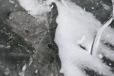 Abstract close-up of frozen lake ice with cracks, trapped air bubbles and a patch of snow, creating...