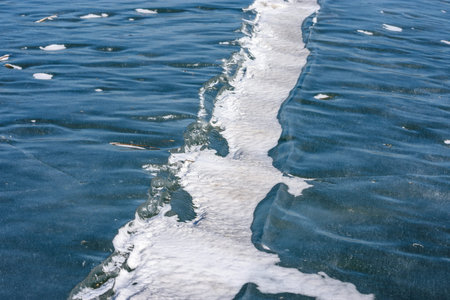 Frozen lake surface with blue ice, scattered snow patches, and intersecting crack lines forming...