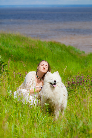 young woman sitting with a big white shaggy dog on a background of the river in the green grassの写真素材