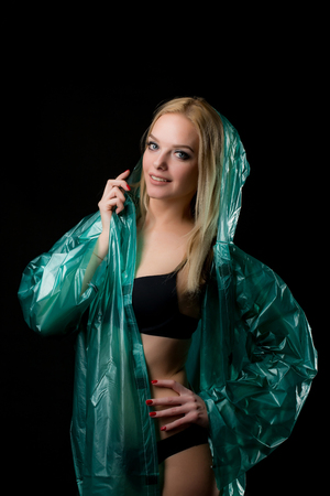 Portrait of a young blond woman in a green raincoat looking at the camera, a hood is put on her head. Shooting in a photo studio on a black backgroundの写真素材