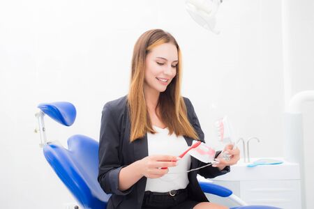 Young beautiful woman in a black jacket shows how to clean teeth with a toothbrush correctly and directly on the jaw model in the dentist office.の写真素材