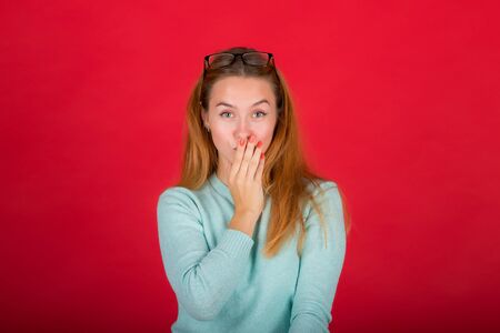 beautiful red-haired girl closes her mouth with her hand, secret, isolated studio photo on a red backgroundの写真素材