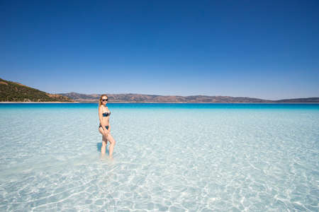 A girl in a bikini swimsuit stands against the background of the water and looks into the distance. Young sexy woman sunbathing in the water of the lake. Beautiful clear blue sky.の写真素材