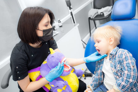 A cute little boy sits at the dentist, opened his mouth, showing his teeth to the dentist. Happy boy is sitting in the dentists chair. Dentist - young brunette womanの写真素材