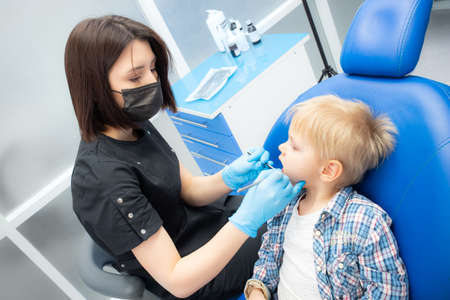 A cute little boy sits at the dentist, opened his mouth, showing his teeth to the dentist. Happy boy is sitting in the dentists chair. Dentist - young brunette womanの写真素材