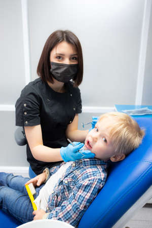 A cute little boy sits at the dentist, opened his mouth, showing his teeth to the dentist. Happy boy is sitting in the dentists chair. Dentist - young brunette womanの写真素材