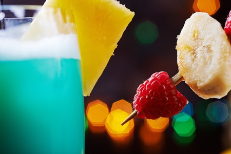 Delicious tropical iced blue curacao alcoholic cocktail garnished with a fresh pineapple slice and maraschino cherries on a table in the restaurant with bright backgrounds of beautiful disco lights. soft focusの写真素材