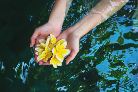 The concept of healthy way of life and purity. Wellness young beautiful girl holding tropical flowers frangipani on clean water in the pool.の写真素材
