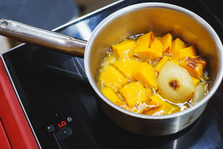 The process of making pumpkin soup - pumpkin sliced into pieces in a saucepan with onion and vegetables. Cooking vegetarian dishes in the kitchen - a pan with vegetables on the hob. Soft focus.の写真素材