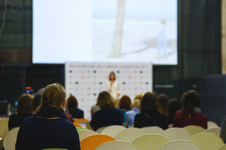 Back view of the participants of the seminar, training or lecture in a large audience with a large screen. The listeners look at the speaker or coach on stage. Beautiful bokeh.の写真素材