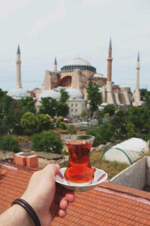 Traditional Turkish tea in a glass on a plate in the hands of a man with a mosque Ayasofya background. Travel to Turkey. Soft focus and beautiful bokeh.の写真素材