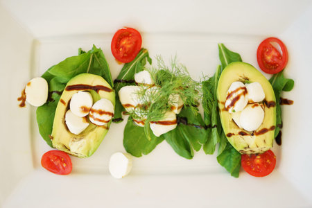 Avocado salad with mozzarella cheese, cherry tomatoes, herbs and sauce in a plate on the table in a restaurant. Delicious and healthy traditional food. Soft focus and beautiful bokeh.の写真素材