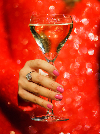 Glass hand girl. White wine in the glass and girl in a sparkling red dress close-up. Celebration and party in a nightclub. Chic red dress. Drinks for adults. Beautiful bokeh and soft focus.の写真素材