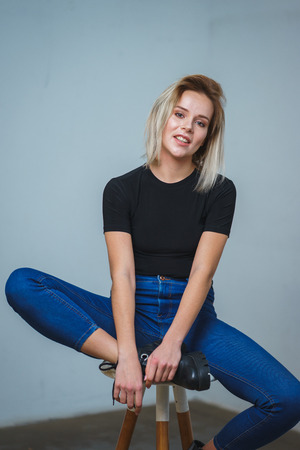 Model tests. Photo session model in studio. Simple attractive portraits for a young beautiful girl in an empty room. Snaps. Attractive girl in jeans,  black shirt. Cheerful girl sitting on a bar stoolの写真素材