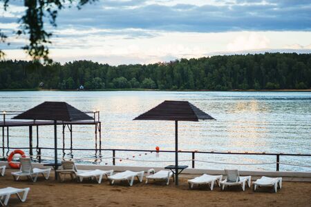 Warm summer evening in city park on the nature shore pond. Lake or river beach from the forest. Summer outdoor recreation. Landscape.の写真素材