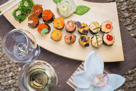 Set of several different rolls on a wooden stand on a table in a Japanese restaurant. Japanese traditional sushi and rolls. Sea products. Beautiful table setting with flowers and wine in glasses.の写真素材
