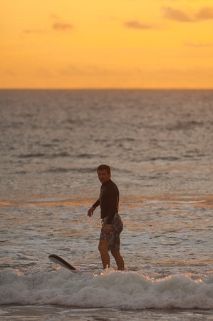 Beginner surfer learns to row and ride on a training board in small waves in the ocean. Golden hour,...