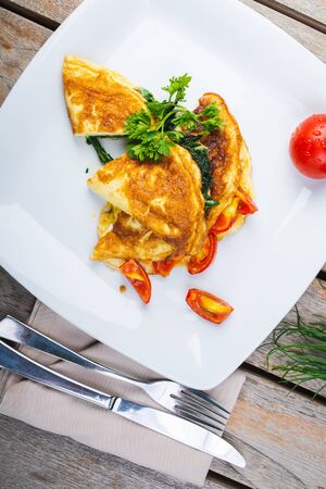 Traditional omelet with vegetables, spinach, tomatoes and herbs on a wooden table in a restaurant. Classic european breakfast. Fried eggs with milk and cheese. Soft focus.の写真素材