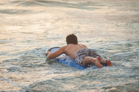 Beginner surfer learns to row and ride on a training board in small waves in the ocean. Golden hour,...