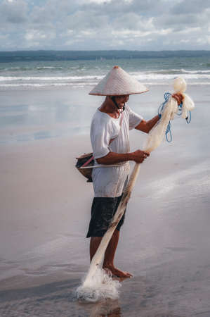 KUTA, BALI - APRIL, 2020: Balinese fisherman with a net and fish catch on the ocean. Travel to Asia. Traditional methods of catching fish by throwing a net. National character. Travel concept.のeditorial素材
