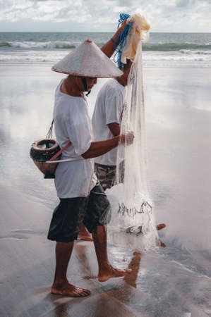 KUTA, BALI - APRIL, 2020: Balinese fisherman with a net and fish catch on the ocean. Travel to Asia. Traditional methods of catching fish by throwing a net. National character. Travel concept.のeditorial素材