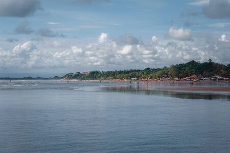Tropical seascape: view of the ocean waves and the beach with tourists relaxing. Sun beds and a parasol on a Balinese beach. Travel to Asia, beach holidays. A warm day by the sea.の写真素材