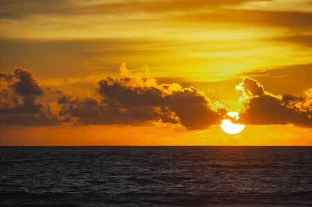 Large bright circle of the sun in the clouds touches the ocean at sunset. Touchdown. Magical dramatic sunset on a tropical beach. Seascape, ocean waves and bright clouds in the sky. Golden hour.の写真素材