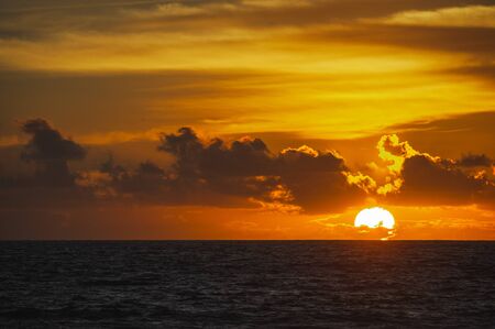 Large bright circle of the sun in the clouds touches the ocean at sunset. Touchdown. Magical dramatic sunset on a tropical beach. Seascape, ocean waves and bright clouds in the sky. Golden hour.の写真素材