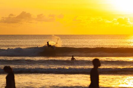 Magical dramatic sunset on a tropical beach. Surfer with a board jumping out of a wave. Seascape, ocean waves and bright clouds in the sky. Golden hour. Rest on the seashore, beach vacation. Concept.の写真素材