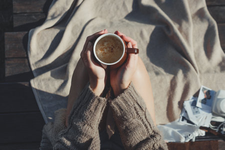 Girl holds a cup of hot coffee in her hands while sitting on the terrace with a plaid on a warm autumn morning. Picnic lawn country house. Autumn mood. Hands Close-up and selective focus.の写真素材
