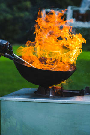 Chef prepares Chinese wok noodles over an open fire on a green lawn. Chefs hands, frying pan and open fire close-up. BBQ with traditional noodles on a summer day.の写真素材