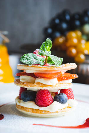 Traditional sweet pastry with three layers of airy dough with whipped cream, with strawberries and berries on a white plate on the table in a cozy cafe with decor. Close-up, macro shot.の写真素材