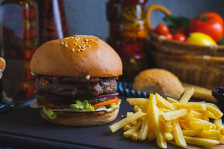 Delicious burger with organic cutlet, cheese, vegetables and fried potatoes on a stone cutting board on wooden table with decor. Fast food in a cafe and restaurant. Menu and content, food.の写真素材