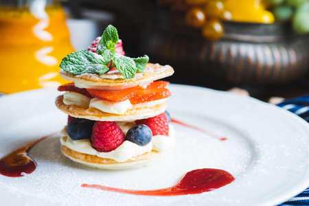 Traditional sweet pastry with three layers of airy dough with whipped cream, with strawberries and berries on a white plate on the table in a cozy cafe with decor. Close-up, macro shot.の写真素材