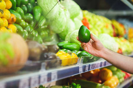 mans hand holds a fresh avocado next to a shelf with vegetables in a supermarket.の写真素材