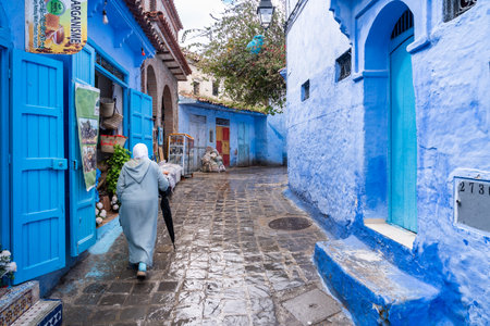 Chefchaouen, Morocco -march 26, 2024:An unrecognizable woman walks with an umbrella among the small shops of the blue city of Chefchaouen, Morocco, on a March day.のeditorial素材
