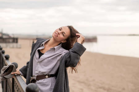 A beautiful young brunette woman walks on the beach in cool weather in a fashionable outfitの写真素材